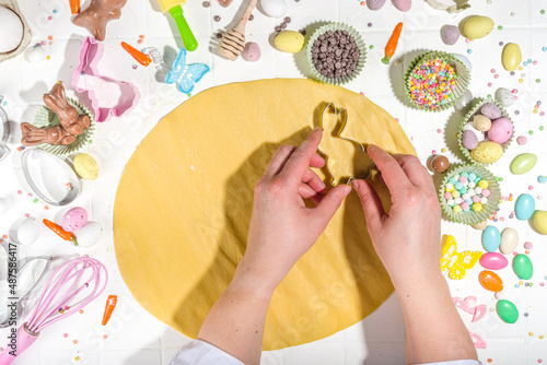 Woman hands kneading dough on table and cooking Easter sugar cookies. Easter baking background with colorful chocolates, candy eggs, baking utencils, ingredients, Top view