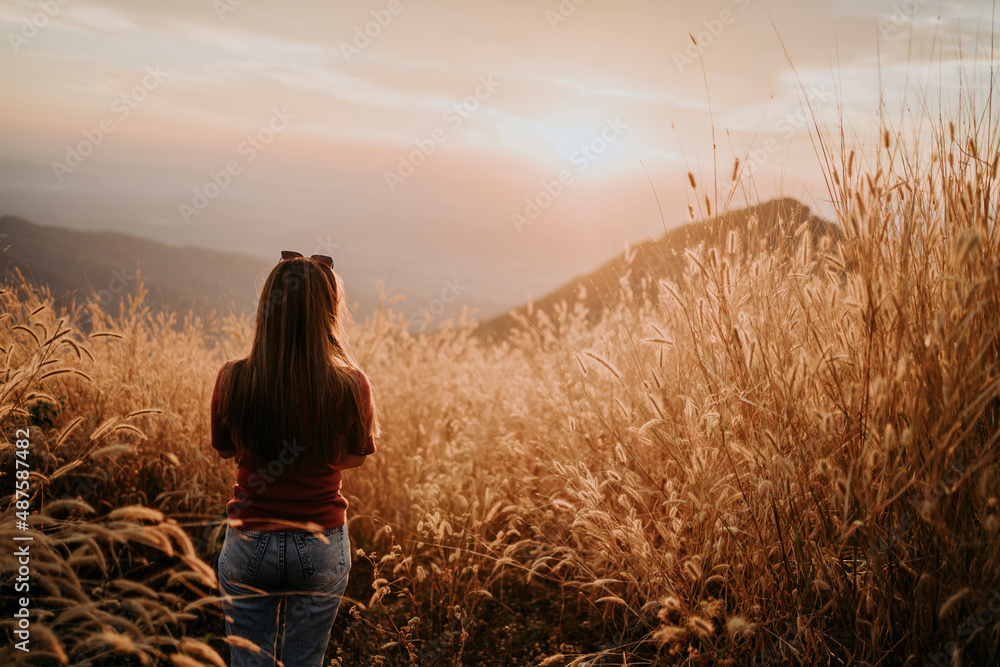 Fototapeta premium A beautiful Asian woman looks at the sunset in the grass in the warm light.
