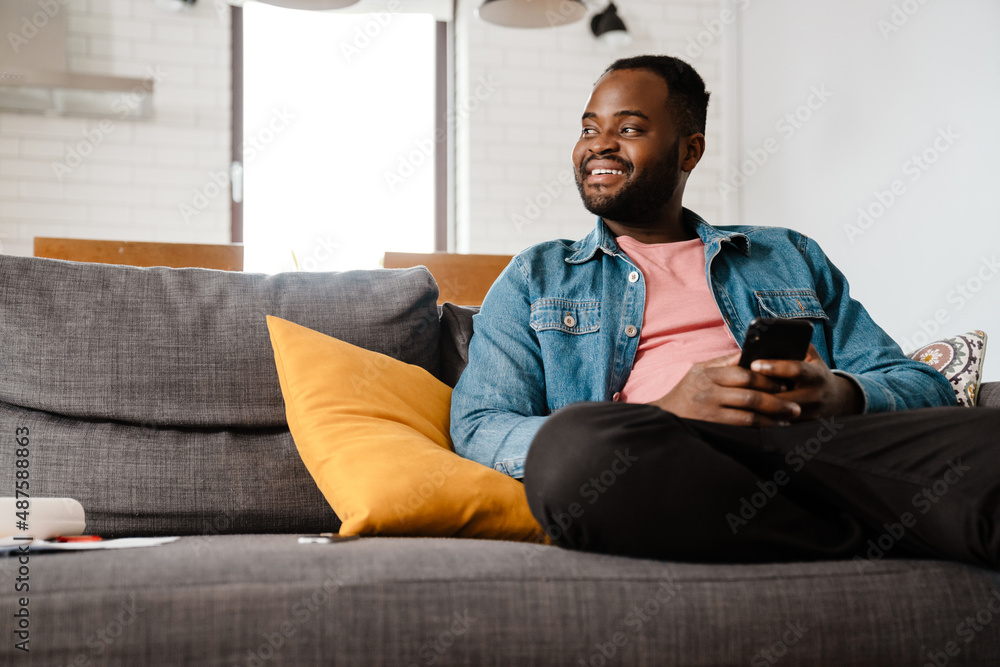 Black bristle man smiling and using cellphone while sitting on sofa Black bristle man smiling and using cellphone while sitting on sofa