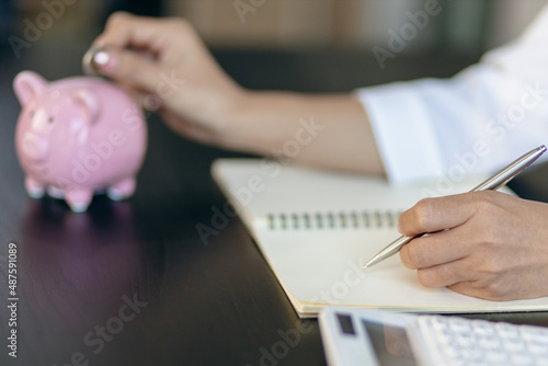 A woman puts coins in a piggy bank and writing a note in the financial book. Money saving concept.