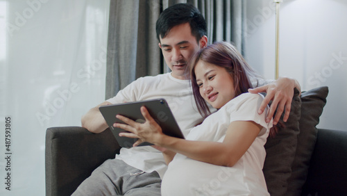 Happy caucasian husband with a pregnant wife looking ultrasound photo of her newborn baby together on a tablet and shopping online in the living room at home.