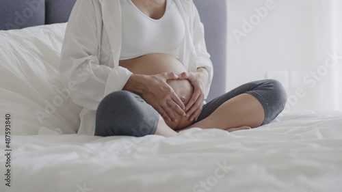 Happy Asian pregnant woman stroking belly while sitting on a bed in the bedroom at home.