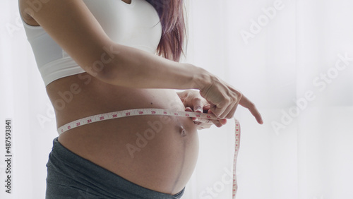 Young Asian Pregnant woman measuring her belly with a tape while standing beside a window in the home.