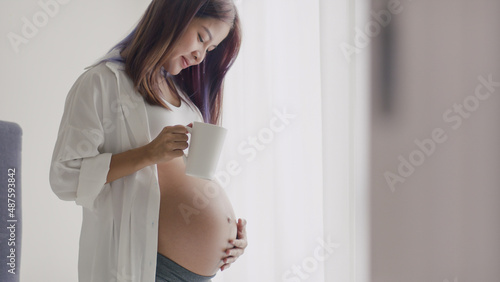 Young Asian Pregnant woman drinking coffee while standing beside a window in the home.