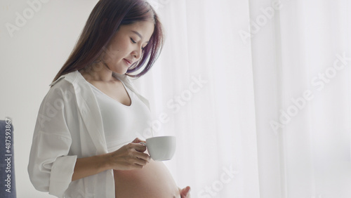 Young Asian Pregnant woman drinking coffee while standing beside a window in the home.