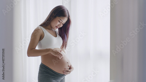 A beautiful Asian happy pregnant woman is standing beside windows and touching her belly.
