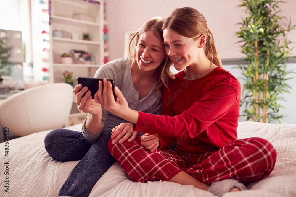 Mother With Teenage Daughter Wearing Pyjamas Sitting On Bed Posing For Selfie On Mobile Phone Mother With Teenage Daughter Wearing Pyjamas Sitting On Bed Posing For Selfie On Mobile Phone