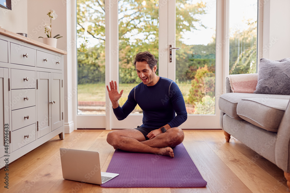 Mature Male Teacher Sitting On Mat At Home With Laptop Giving Online Yoga Class Mature Male Teacher Sitting On Mat At Home With Laptop Giving Online Yoga Class