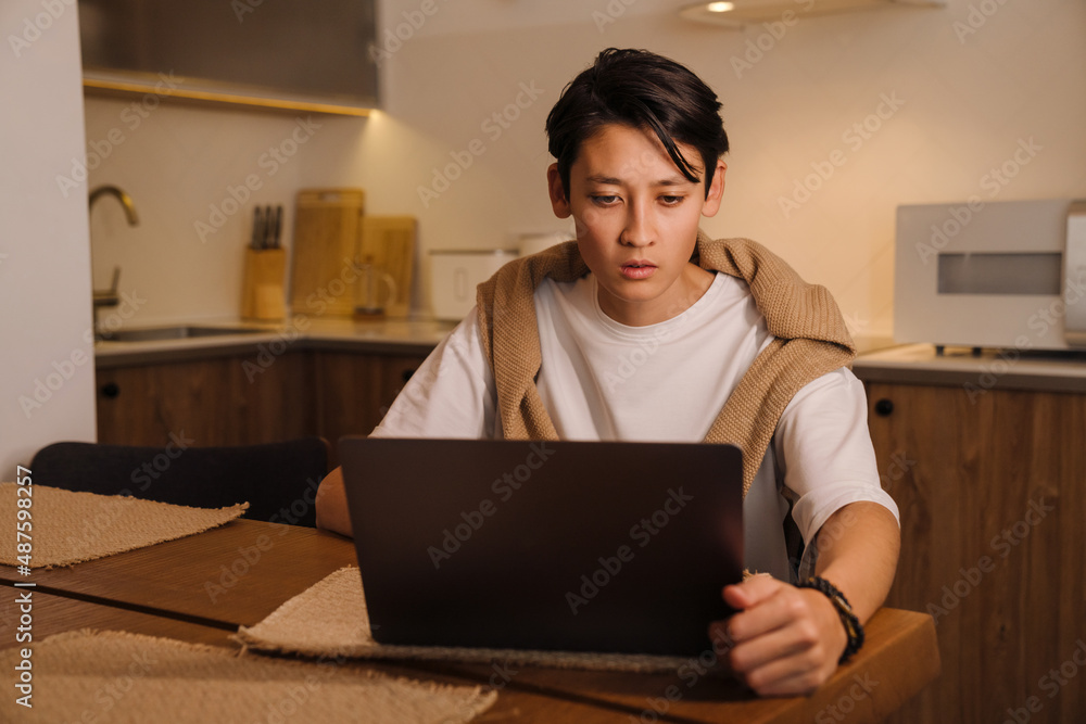 Asian boy working with laptop while sitting in kitchen Asian boy working with laptop while sitting in kitchen