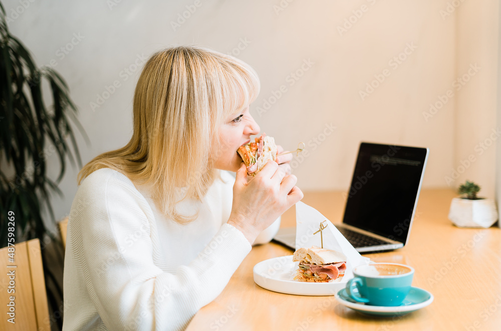 Mature adult woman sitting in cafe with coffee mug eating sandwiches and working online at laptop. Businesswoman in typing on notebook computer, in coworking space in roasters coffee shop. Mature adult woman sitting in cafe with coffee mug eating sandwiches and working online at laptop. Businesswoman in typing on notebook computer, in coworking space in roasters coffee shop.