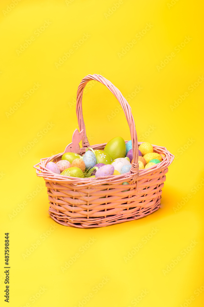 Easter basket full of colorful Easter eggs, on a yellow background. Easter holiday concept, photo with a copy space.