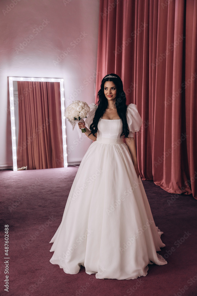 The bride in a white dress in front of the mirrors in the living room. Preparing for the upcoming wedding. Burgundy background. The bride in a white dress in front of the mirrors in the living room. Preparing for the upcoming wedding. Burgundy background.