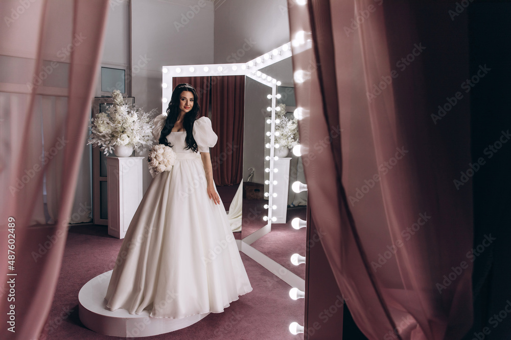 The bride in a white dress in front of the mirrors in the living room. Preparing for the upcoming wedding. Burgundy background. The bride in a white dress in front of the mirrors in the living room. Preparing for the upcoming wedding. Burgundy background.