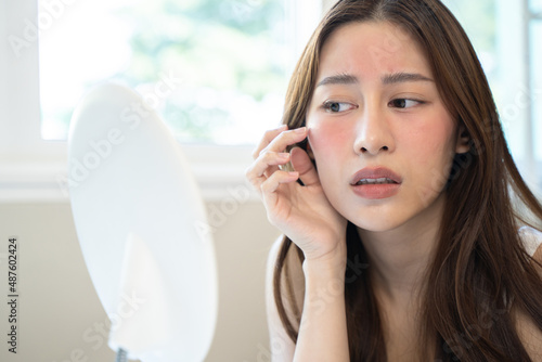 Asian young woman looking face skin in the mirror have a red rash on her face from cosmetic allergy