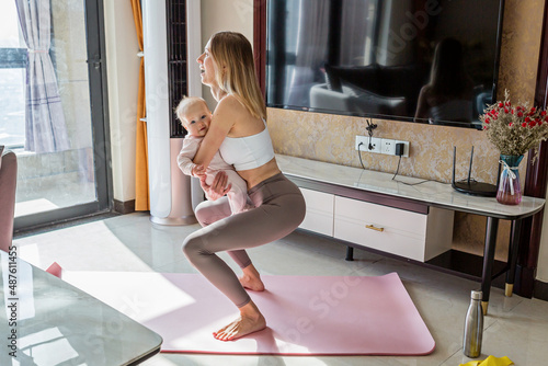Young mother in sport clothing exercising at home with baby. Online training during coronavirus covid-19 quarantine. Stay fit and safe during pandemic lockdown. Sport, fitness, healthy concept
