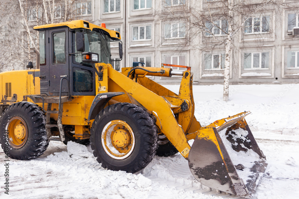 Big orange tractor cleans up snow from the road and loads it into the truck. Cleaning and cleaning of roads in the city from snow in winter. Snow removal after snowfall and blizzards. 