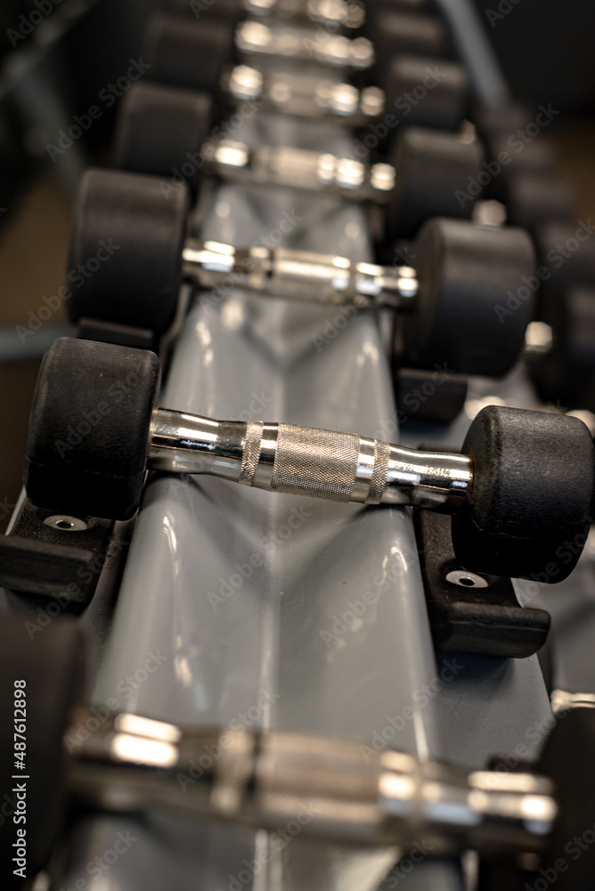 Dumbbells with silver handles and black rubber end plates on a rack, shallow depth of field. Dumbbells with silver handles and black rubber end plates on a rack, shallow depth of field.
