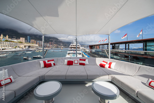 The front deck of huge yacht in port of Monaco at sunny day, landmarks of Monte-Carlo and a lot of motorboats are on background, the lounge zone for a chilling, megayacht is moored in marina