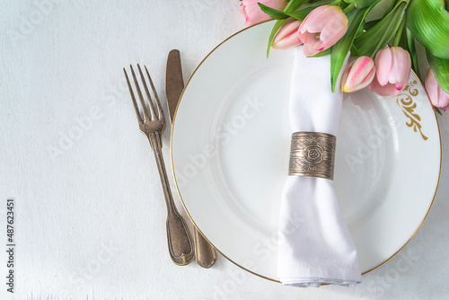 Spring table setting; Bouquet of pink tulips, vintage fork and knife, white napkin with ring on white plate top view