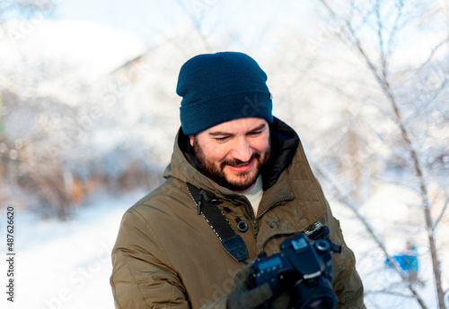 A bearded man in a winter hat and clothes holds a camera in mittens, looking at his screen. Photographer during photo shoot in winter outdoors © PeterPike