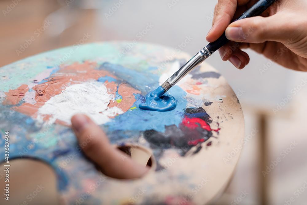 cropped view of woman mixing colors on palette with paintbrush.