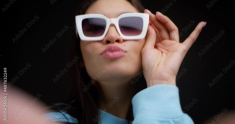 Close-up of the face of a young beautiful brunette in white glasses taking a selfie on a smartphone. Young woman in blue clothes takes pictures of herself on her phone on a black background. Close-up of the face of a young beautiful brunette in white glasses taking a selfie on a smartphone. Young woman in blue clothes takes pictures of herself on her phone on a black background.