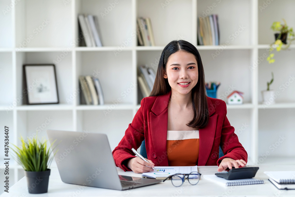 Confident beautiful Asian businesswoman using a calculator and taking notes in office. Looking at camera. Confident beautiful Asian businesswoman using a calculator and taking notes in office. Looking at camera.