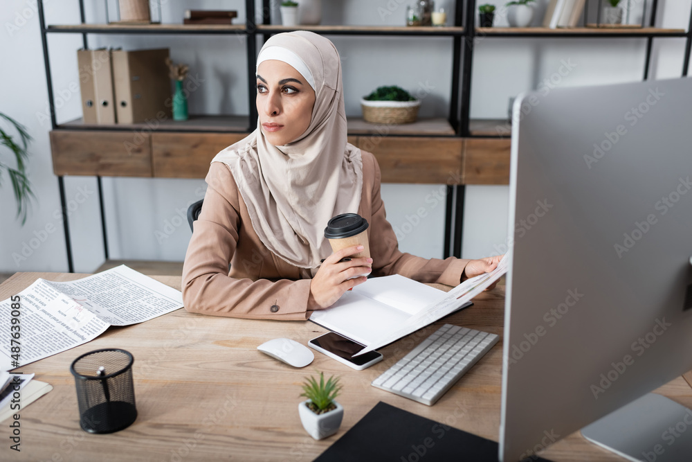 muslim woman looking away and holding coffee to go while sitting in home office. muslim woman looking away and holding coffee to go while sitting in home office.