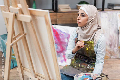 muslim woman with palette and paintbrushes looking at canvas at home.