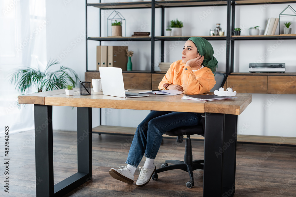 dreamy muslim woman looking away while sitting at desk near laptop.