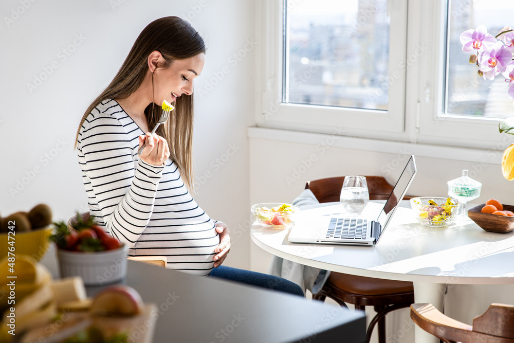 Pregnant woman sitting at table at home eating fruit salad and using laptop.