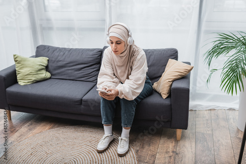young muslim woman sitting on couch with smartphone and listening music in headphones. © LIGHTFIELD STUDIOS