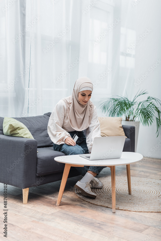 muslim woman sitting on sofa with credit card near laptop on coffee table.