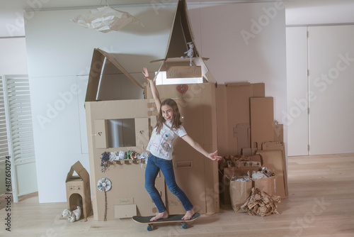 Kid on a skateboard, indoors, playing, a home-made cardboard house in the background
