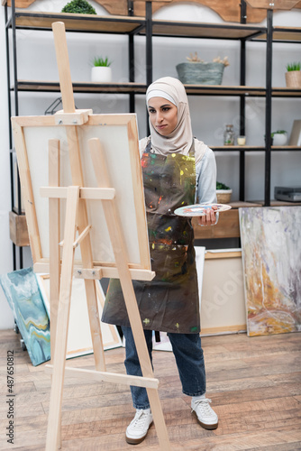 full length view of muslim woman painting on easel in home studio.