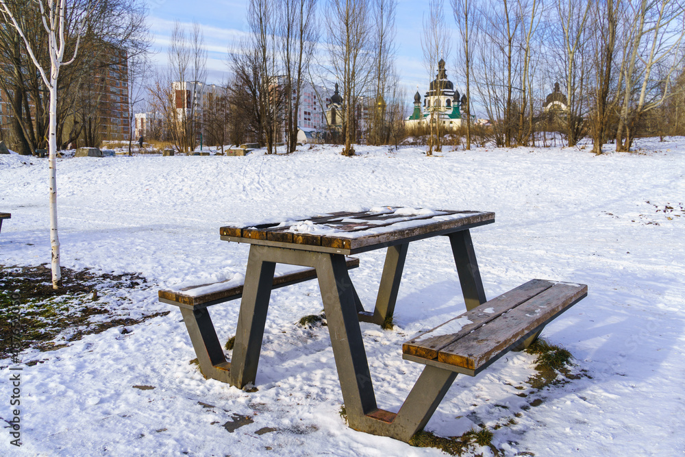 Wooden table and benches for a picnic on a winter snowy day against the backdrop of a Christian temple, city houses and blue sky