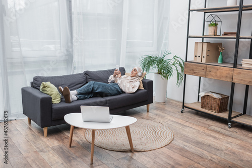 happy muslim woman listening music in headphones while lying on couch in living room.