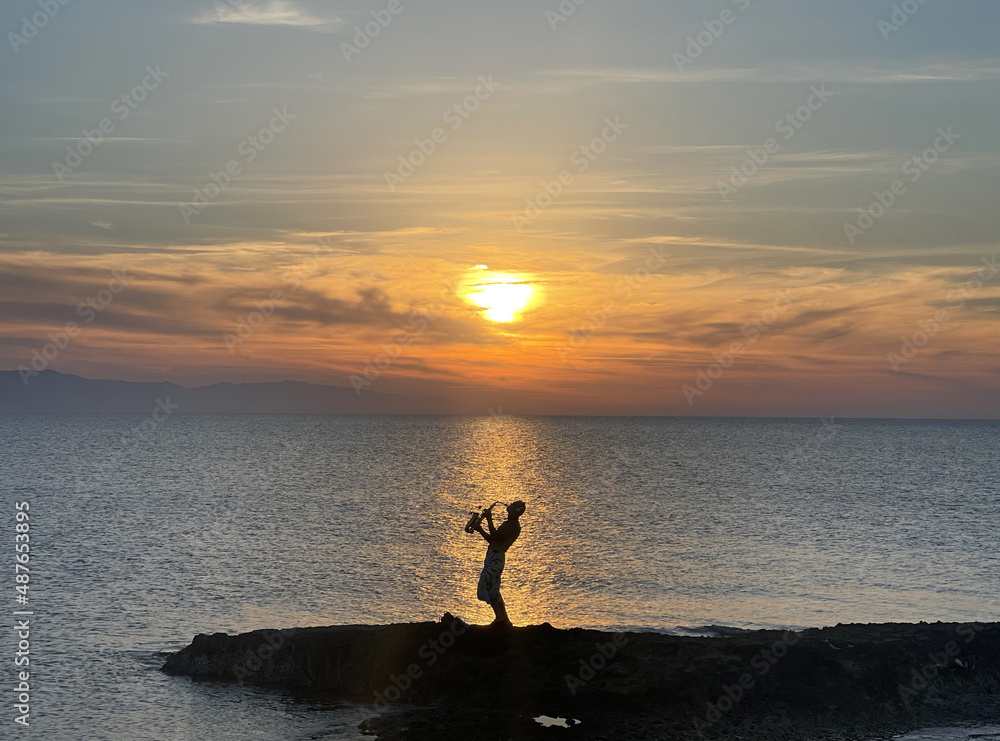Young male saxophonist stands against background of sea and sunset, holds saxophone in his hands. Beautiful sunset on sea, sky. Musician saxophonist plays against background sea, sunset, silhouette