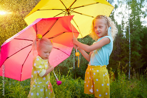 Cute little blonde girls with umbrella under rain drops on lawn in a sunny summer day. Two small friends having rest together