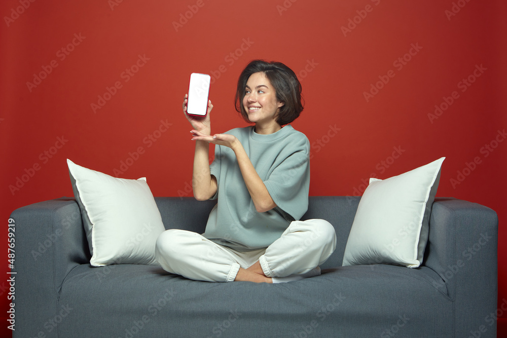 Smiling beautiful young girl showing phone with blank white screen, sitting on sofa. Modern mobile apps advertising Smiling beautiful young girl showing phone with blank white screen, sitting on sofa. Modern mobile apps advertising