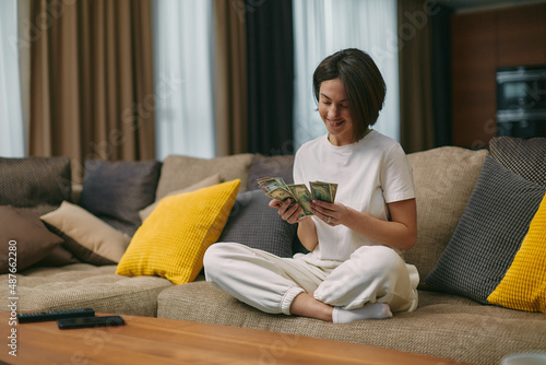 Happy young woman counting cash, enjoying money win, big profit, salary in payday sitting on sofa at home photo