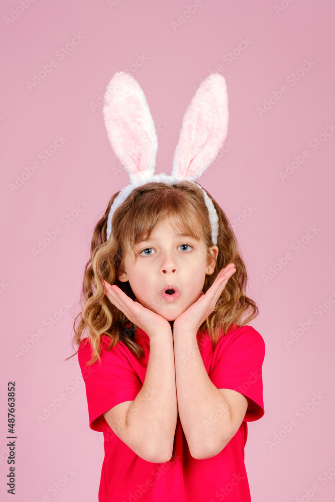 Adorable surprised little girl with long curly hair in Easter bunny ears touching cheeks and looking at camera with open-eyes, against pink background in studio, vertical orientation