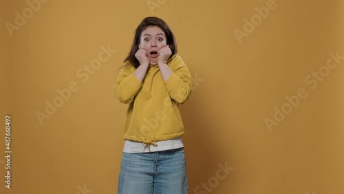 Portrait of woman being scared doing hand gesture touching face in fear feeling frightened and afraid over yellow background. Young person doing shocked face grimace being startled in studio.