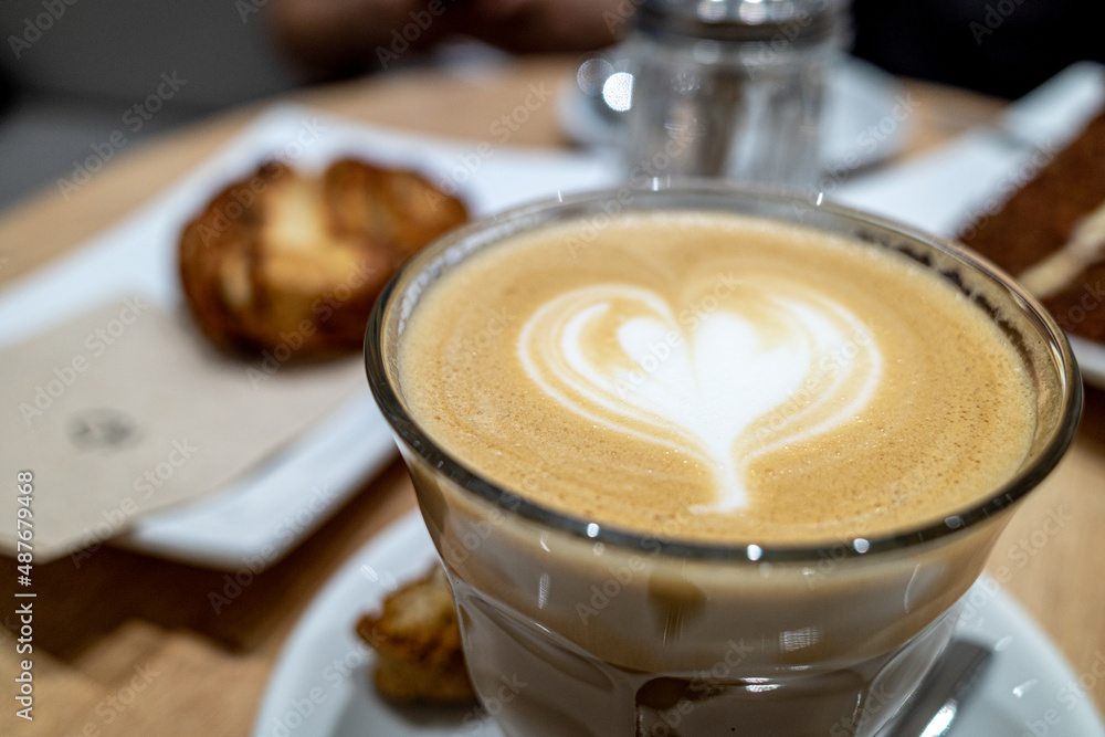 Selected focus and close up view of a white ceramic cup of cappuccino on top with beautiful delicate latte art with flower and heart pattern, on wooden outdoor counter of coffee shop. Selected focus and close up view of a white ceramic cup of cappuccino on top with beautiful delicate latte art with flower and heart pattern, on wooden outdoor counter of coffee shop.