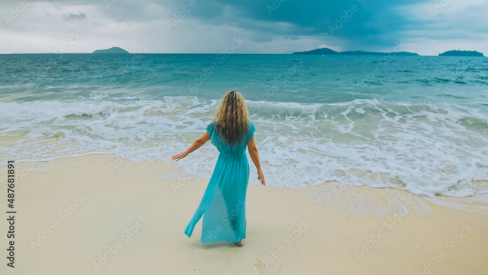 Charming woman with blonde curly long hair walk in beach, in stormy morning rain cloudy sea. Girl in turquoise swimsuit tunic. Concept resort outdoor relax summer vacation, travel. Dark dramatic view Charming woman with blonde curly long hair walk in beach, in stormy morning rain cloudy sea. Girl in turquoise swimsuit tunic. Concept resort outdoor relax summer vacation, travel. Dark dramatic view