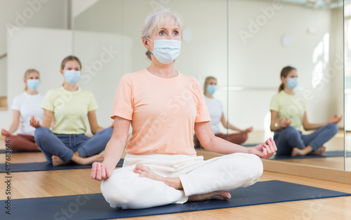 Senior woman in face mask sitting on mat in lotus pose during group yoga training with her family.