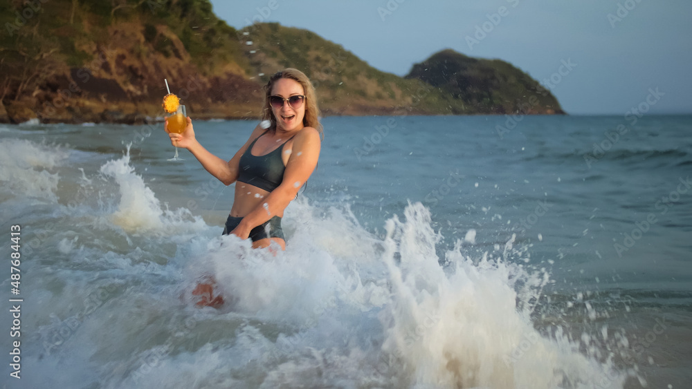 Sexy hot woman stand knee-deep in sea on golden sunset. Girl on tropical beach in green swimsuit having fun and waving his hands, drinks her orange cocktail Pina Colada. Big waves hitting buttocks