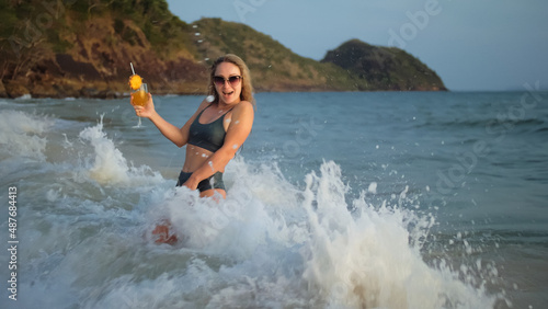 Sexy hot woman stand knee-deep in sea on golden sunset. Girl on tropical beach in green swimsuit having fun and waving his hands, drinks her orange cocktail Pina Colada. Big waves hitting buttocks