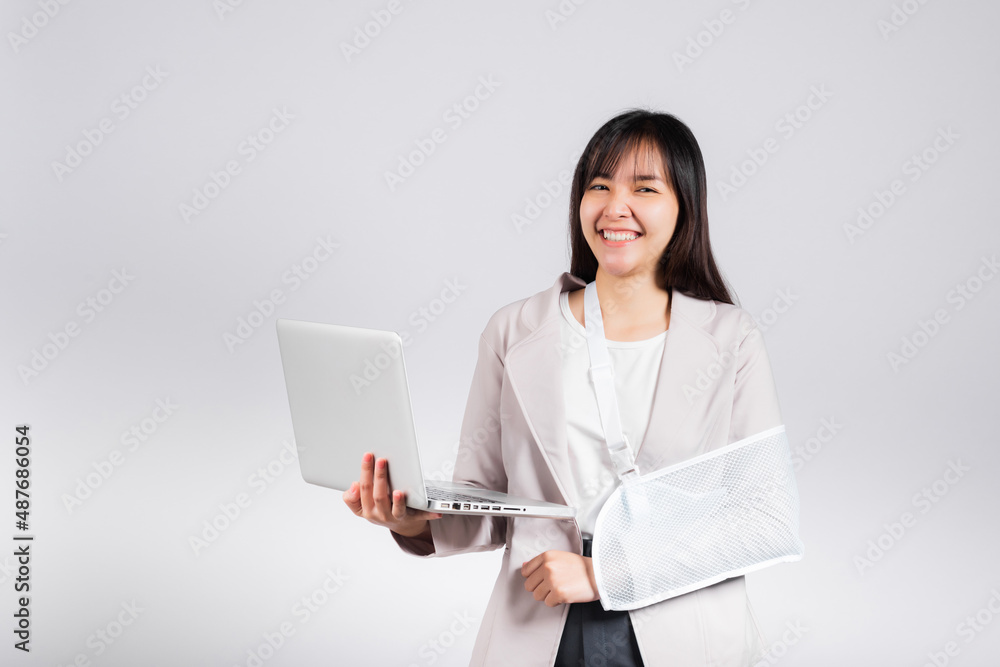Asian business woman confident smiling broken arm after accident and wear arm splint for treatment but still have to work she hold laptop computer studio isolated on white background,copy space Asian business woman confident smiling broken arm after accident and wear arm splint for treatment but still have to work she hold laptop computer studio isolated on white background,copy space