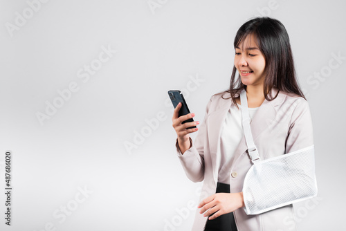 Woman confident smiling broken arm after accident and wear arm splint for treatment and hold smartphone, happy Asian female sling supported hand isolated on white background, social media, copy space photo